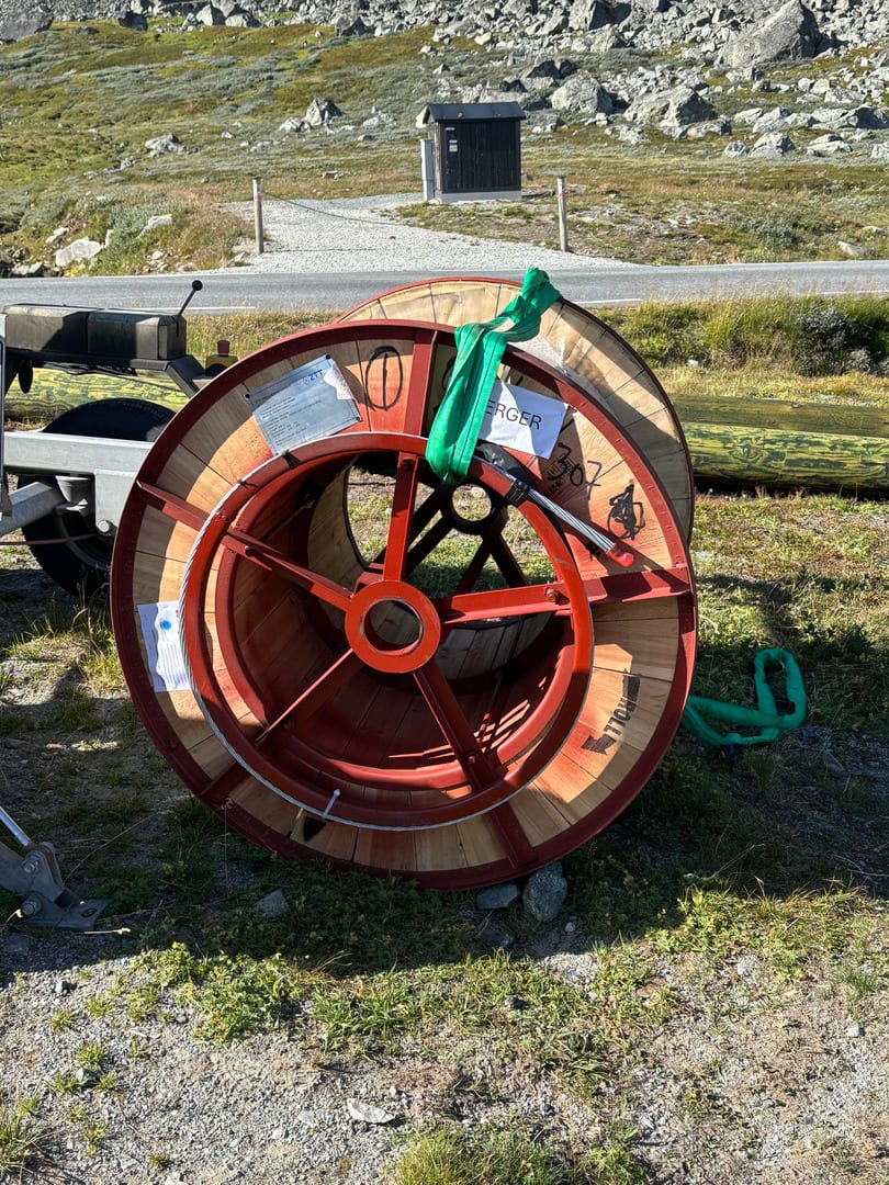 Stor kabeltrommel med metallramme og trekjerne ligger på bakken ved fjellvei, klar for bruk i strømprosjektet.