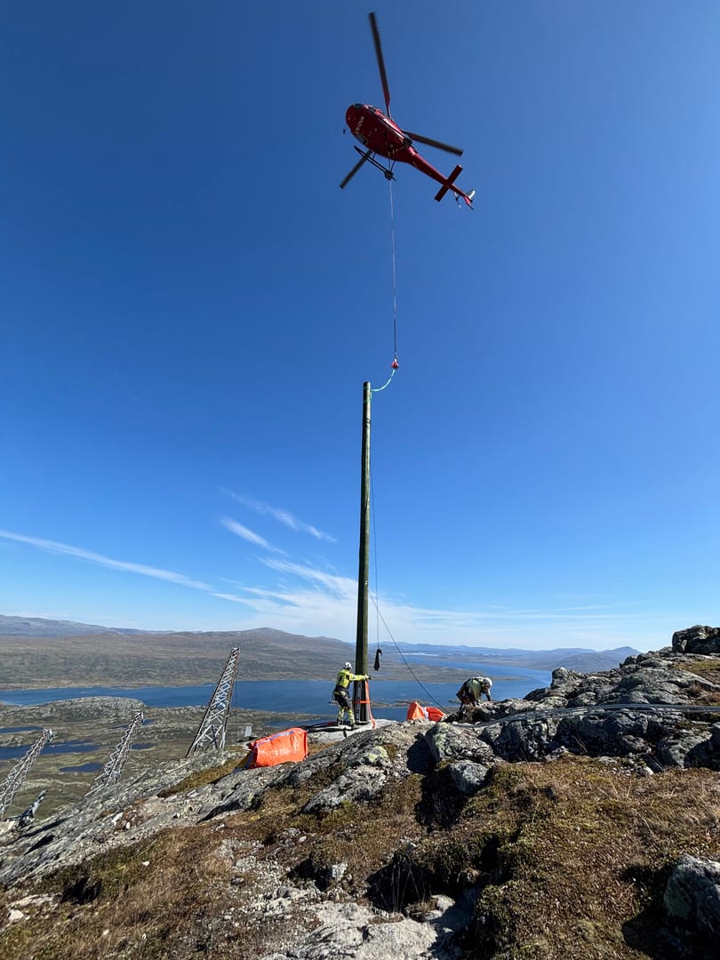 Et helikopter løfter en stolpe på plass mens to linjearbeidere sikrer den på fjelltoppen under klar blå himmel, med innsjøer og fjell i bakgrunnen.