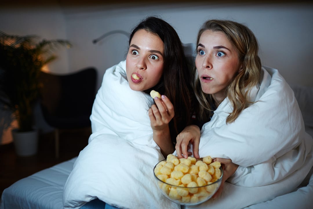 To personer sitter under et hvitt teppe med en bolle snacks og følger intenst med på noe på skjermen foran seg. Scenen formidler en koselig filmkveld hjemme – et øyeblikk av avslapning og underholdning.