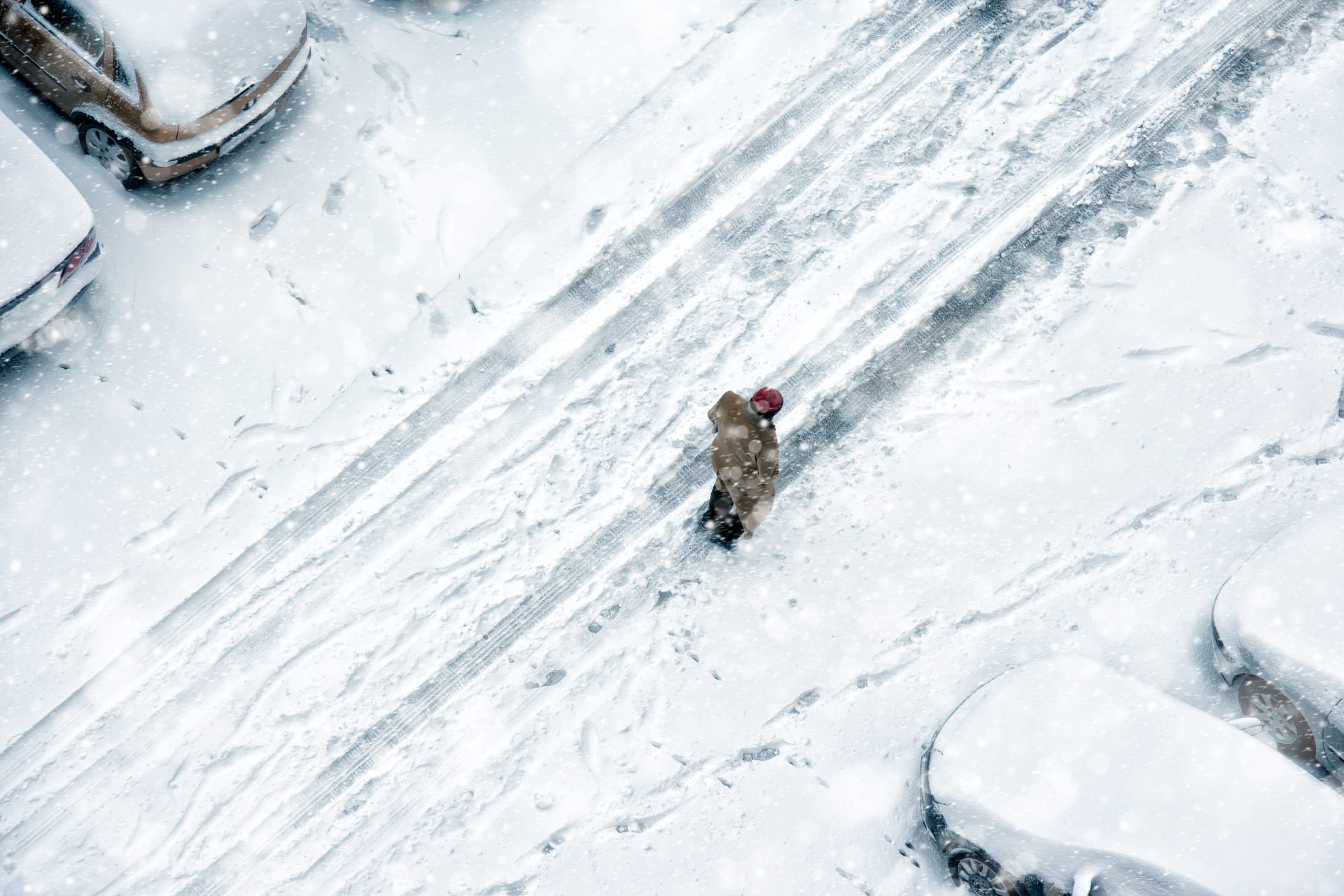 Aerial view of a snowy street with parked cars, a person walking, and falling snow.